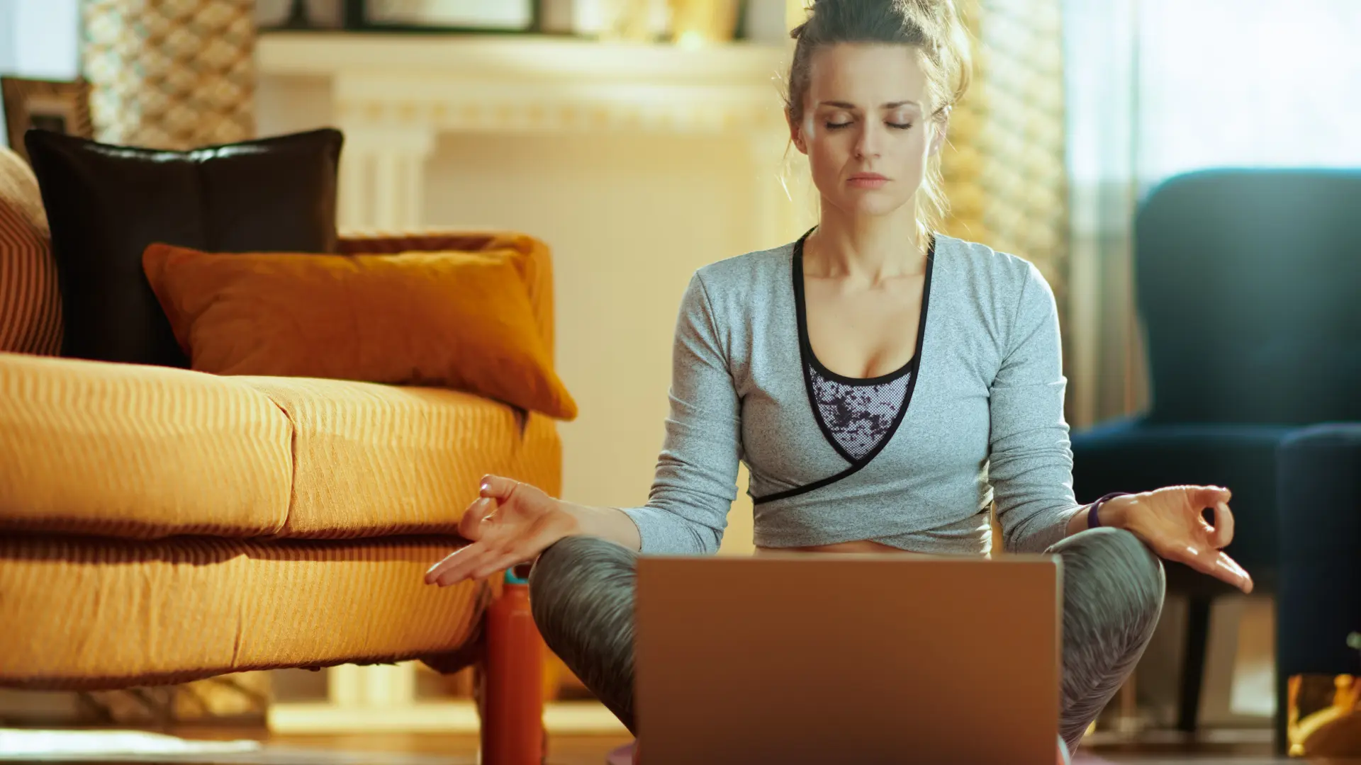 Frau in einem Wohnzimmer in Meditationshaltung vor einem Laptop am Meditieren.