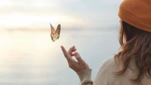 Frau mit gelber Wollkappe, die vor einem See steht und den Finger ausstreckt und ein Schmetterling fliegt in der Luft
