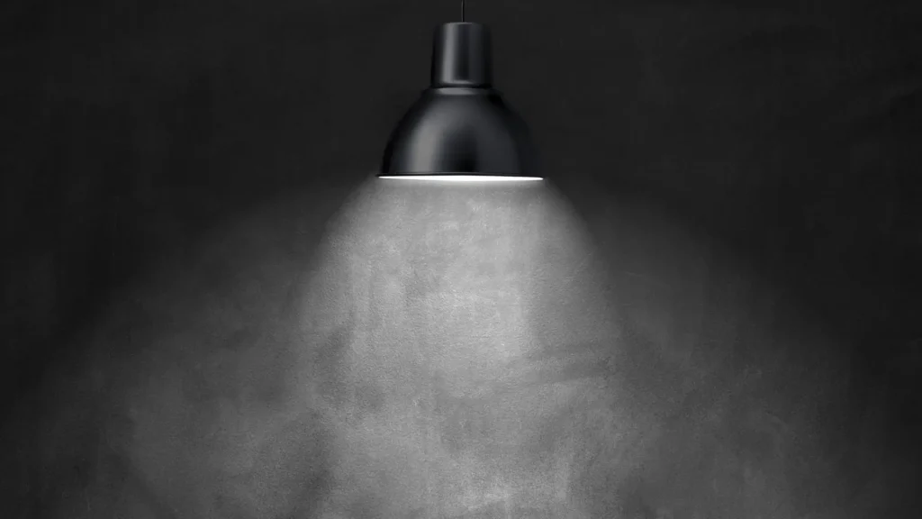 Schwarz-weiße Darstellung einer Lampe, die in einem dunklen Raum einen Lichtkegel auf eine Wand wirft – Symbolbild für Gaslighting und das Infragestellen der eigenen Realität