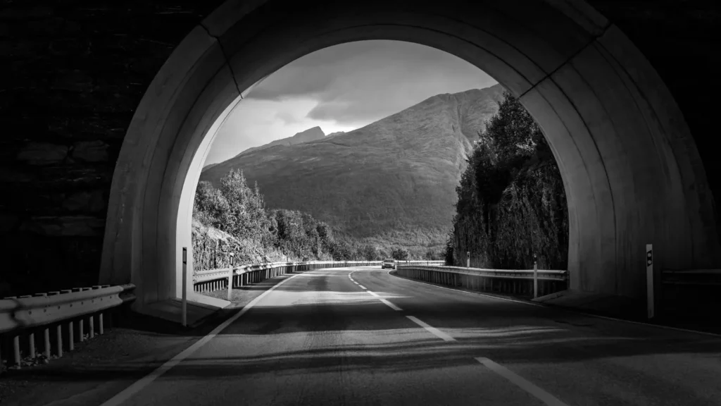 Schwarz-weißes Foto einer Straße, die durch einen Tunnel führt und sich in eine offene Berglandschaft öffnet – Symbol für Übergang, Orientierung und das Verlassen eines Schutzraums in Richtung Weite und Sicherheit.