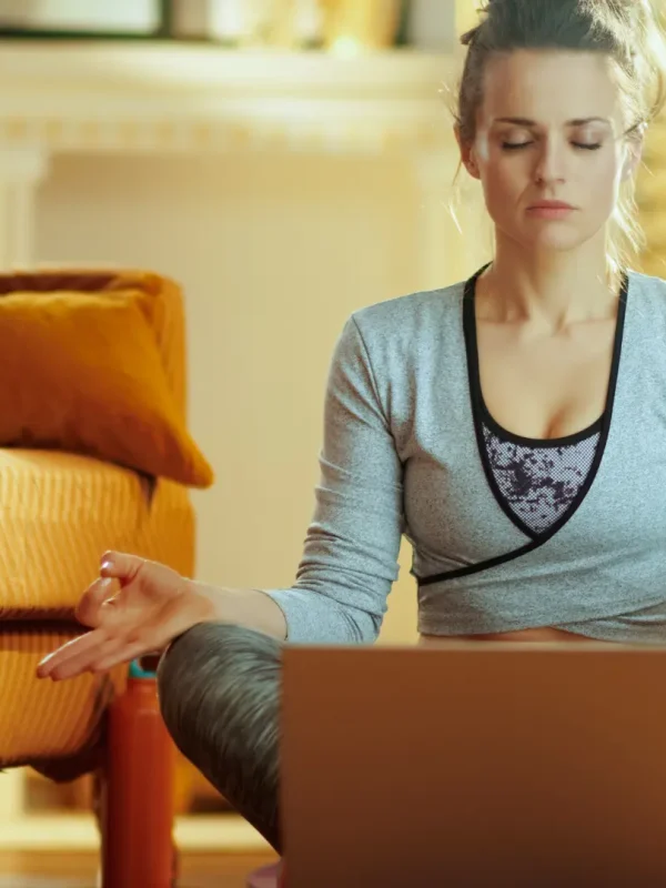 Frau in einem Wohnzimmer in Meditationshaltung vor einem Laptop am Meditieren.